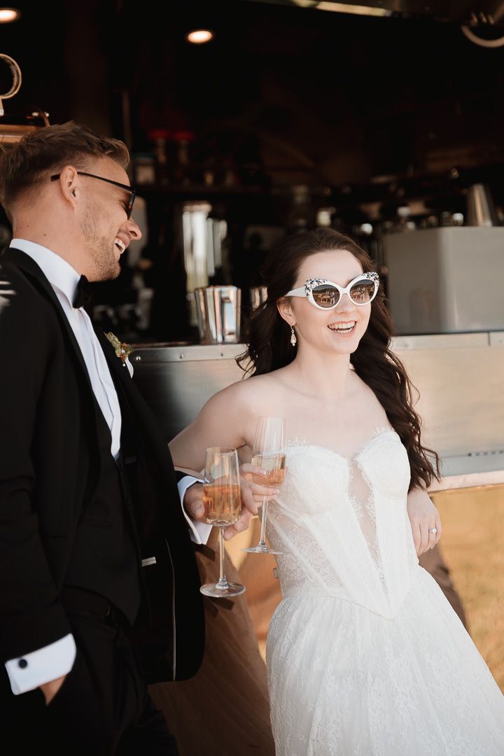A bride in a strapless white dress wearing retro sunglasses laughs while standing at a bar counter.