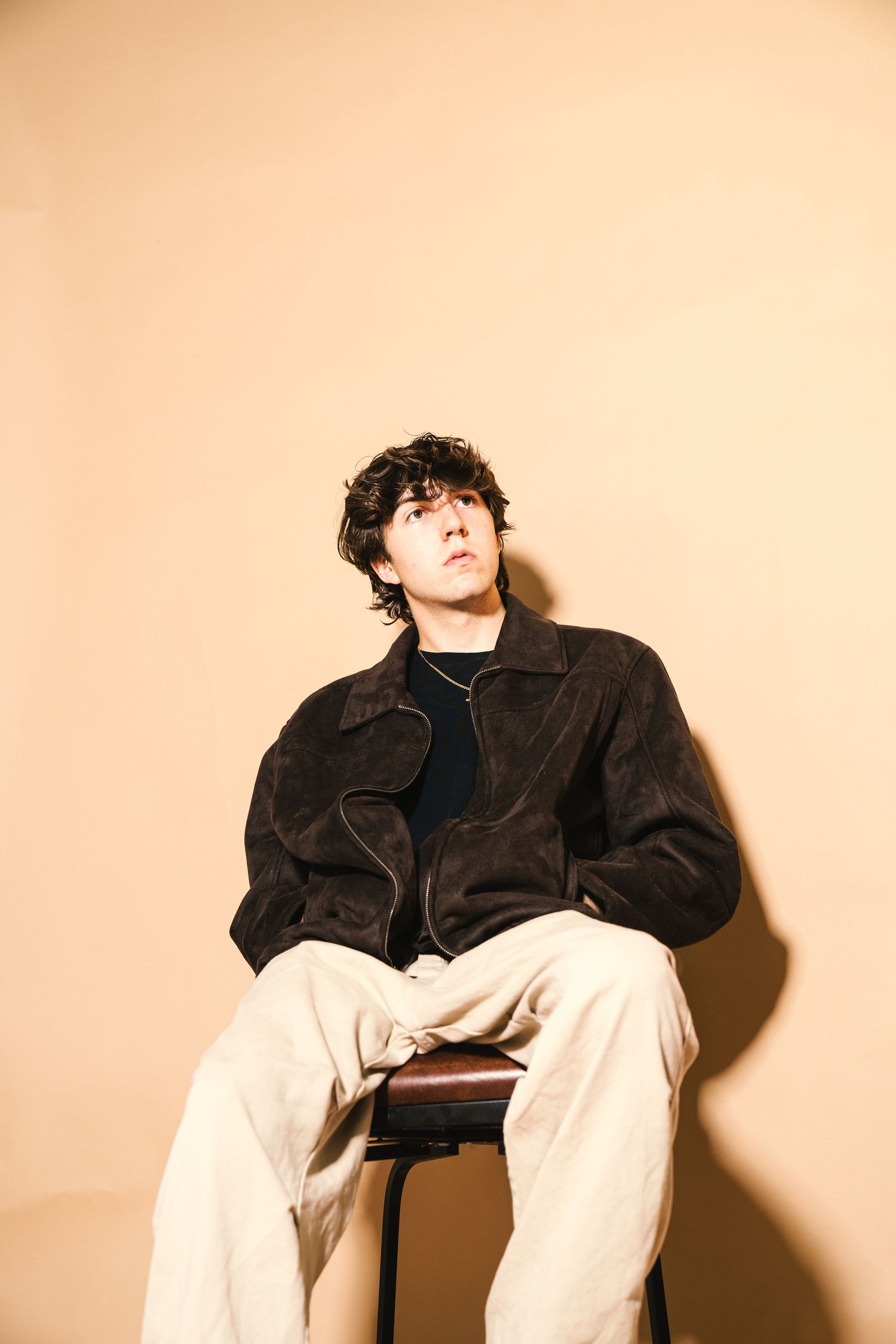 Artistic portrait of person in black leather jacket seated against beige background.