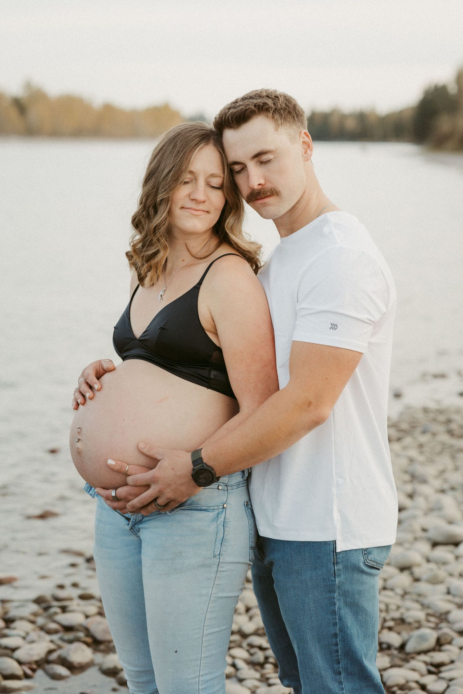 Couple embraces on a rocky beach shore during sunset maternity photoshoot.