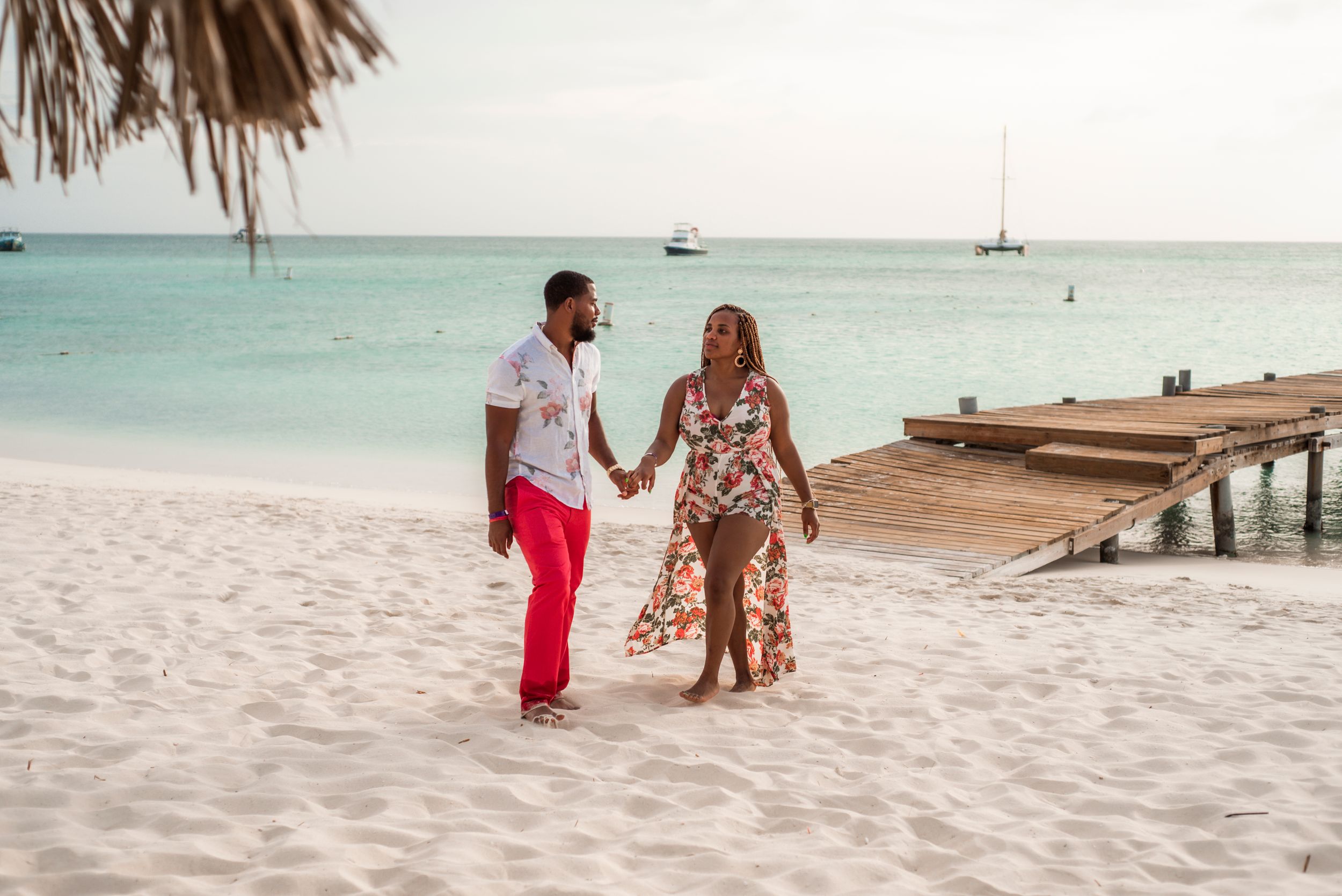 Couple walking hand in hand on a tropical beach with turquoise waters and wooden pier in the background.