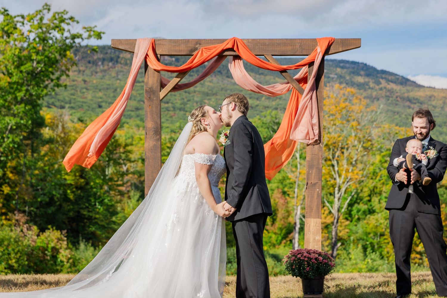 Rustic outdoor wedding ceremony under wooden arch with flowing coral fabric and mountain views.