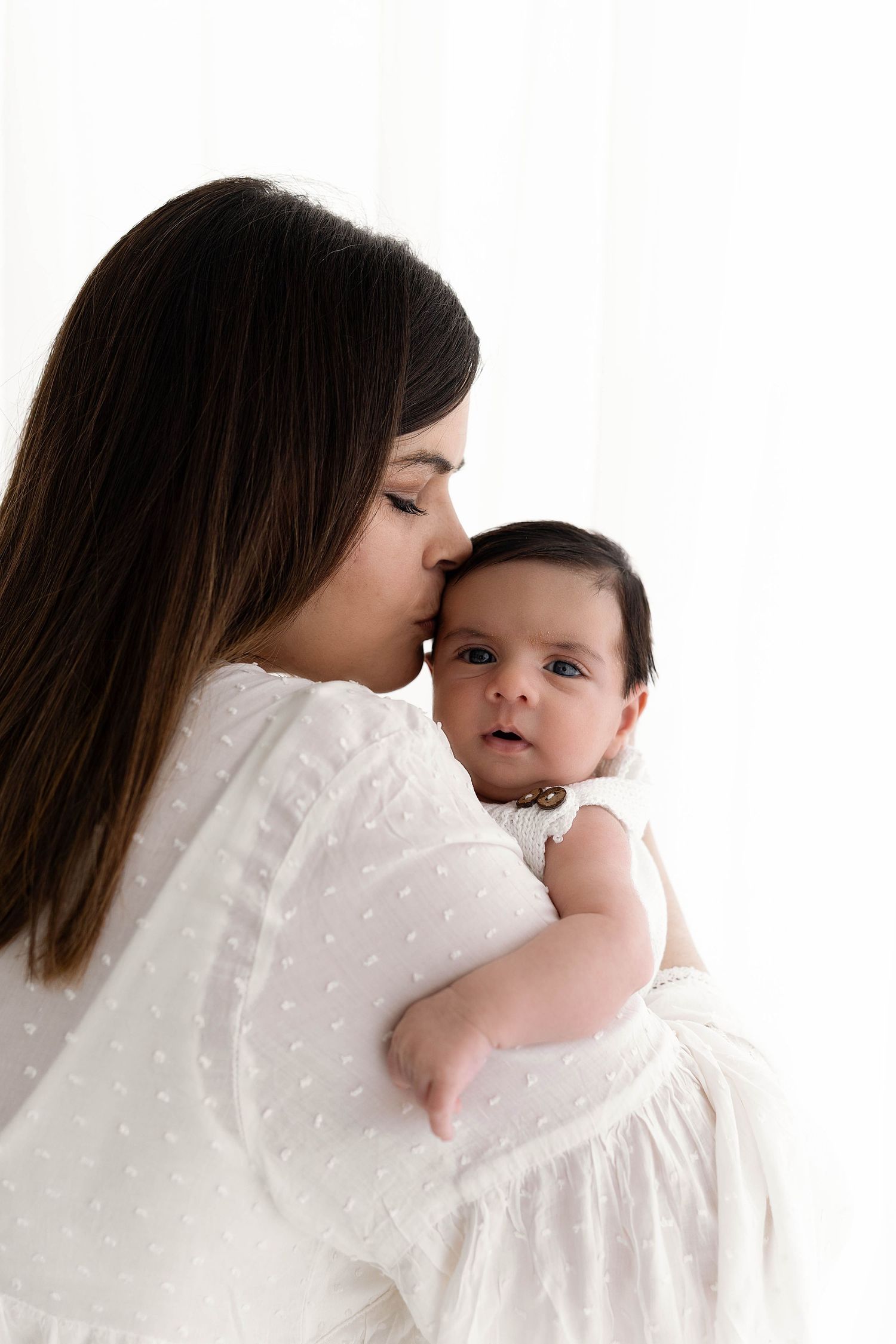 Mother in white dotted dress snuggles with baby against white background.