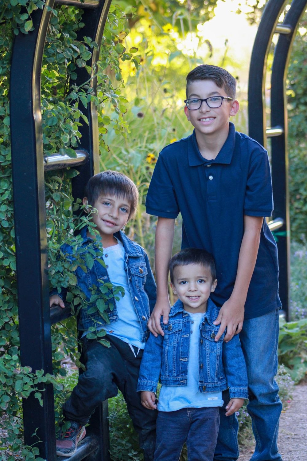 Three children pose together under garden arch with climbing vines.