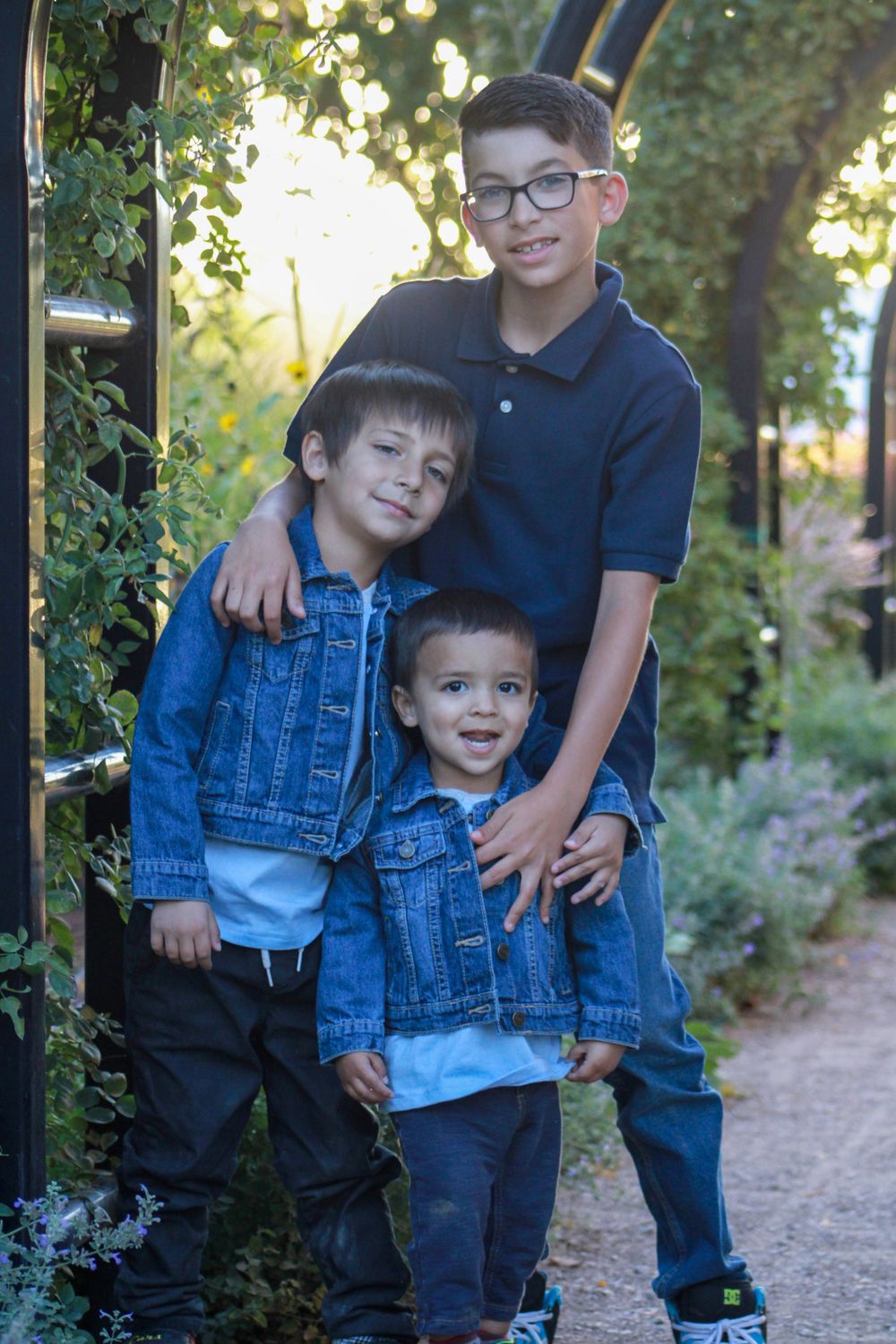Three siblings in coordinating outfits pose together under garden arbor.
