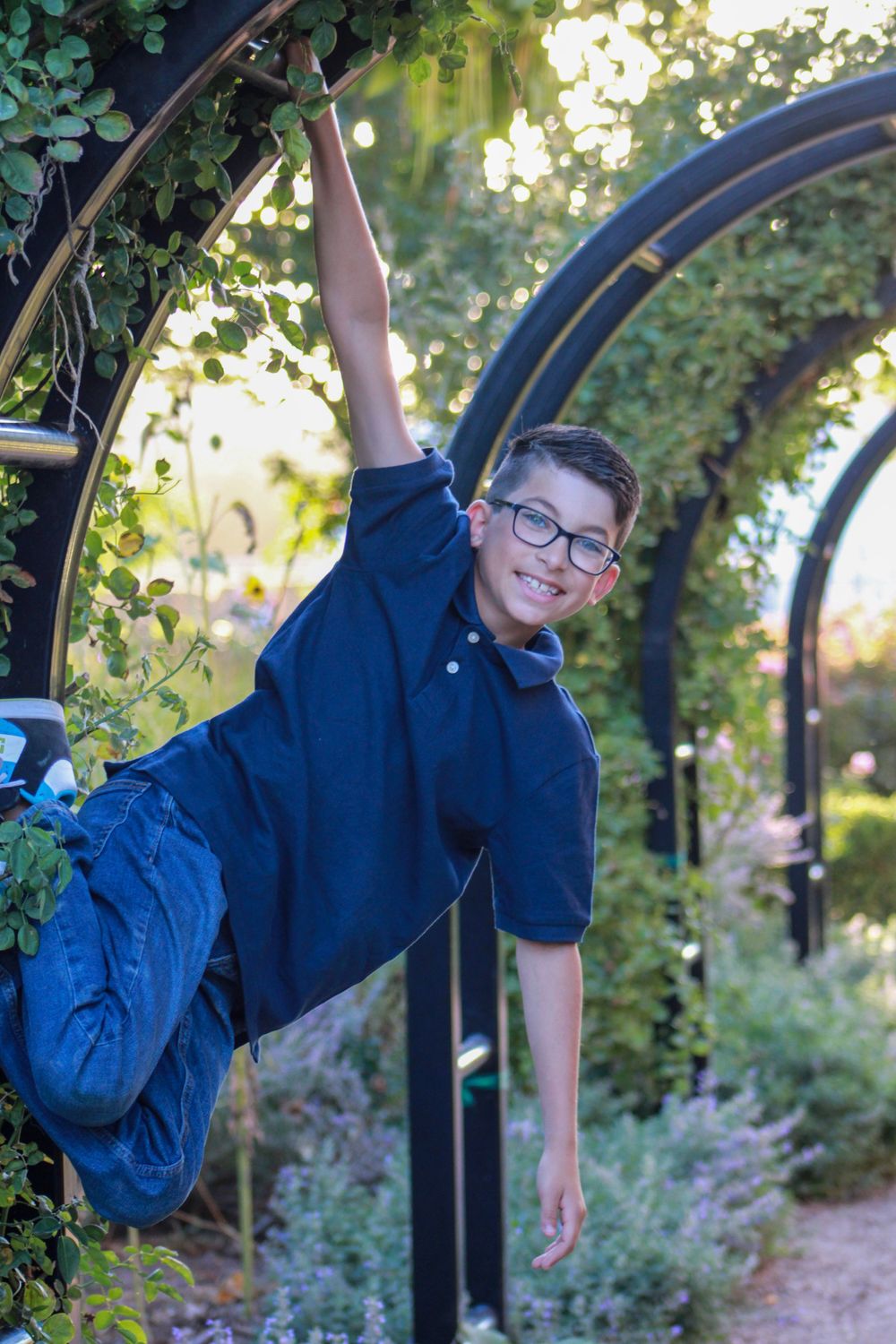 Child hanging playfully from a garden arch covered in greenery during golden hour.