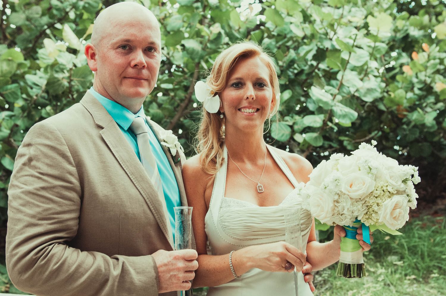 A couple in wedding attire poses outdoors, with the bride holding a white bouquet against a leafy green backdrop.