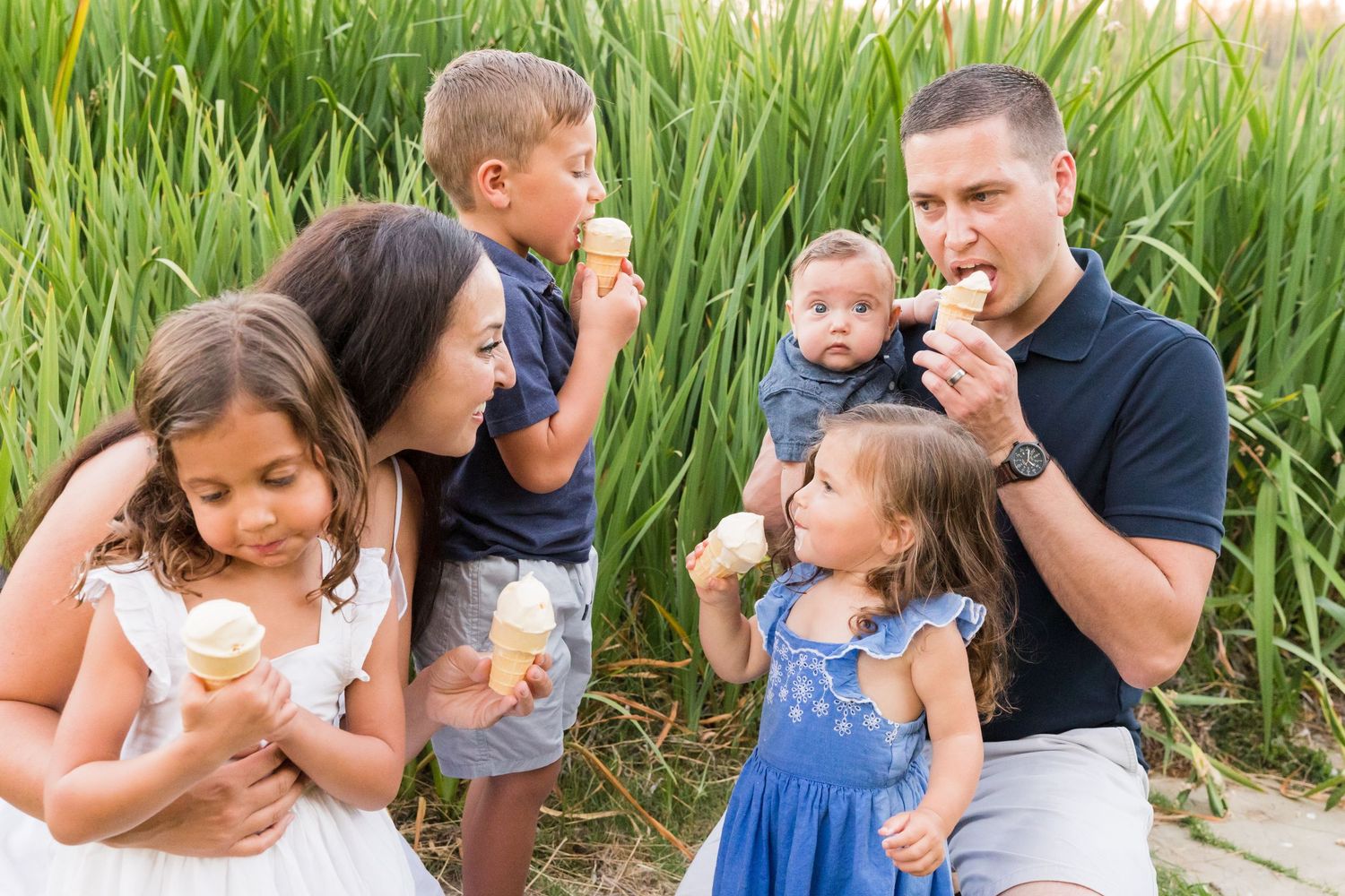 Family of six enjoying ice cream cones outdoors near tall green grass on a sunny day.