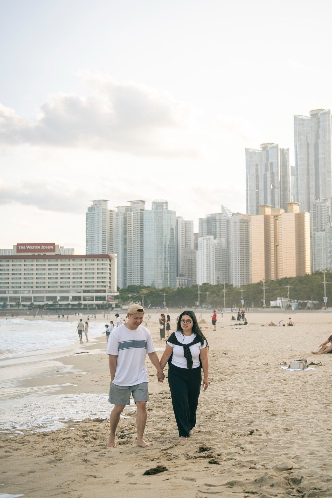 Couple holding hands walking on Haeundae Beach in Busan, South Korea, with city skyline in background.