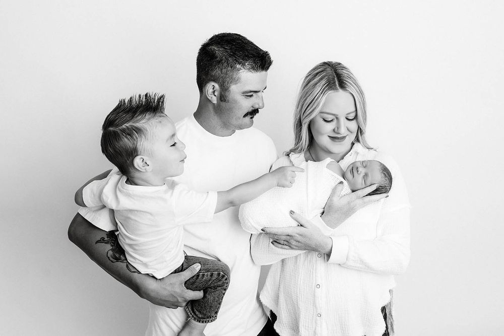 Black and white family photo of parents with toddler son and newborn baby in a bright studio setting by Romoland newborn photographer.