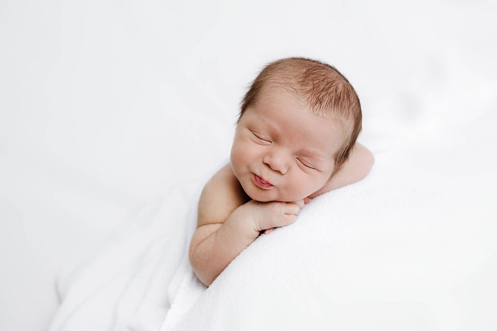 Peaceful newborn baby sleeping wrapped in white blanket with a gentle smile by Romoland newborn photographer.