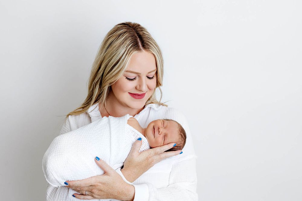 Smiling blonde woman in white outfit gently cradling a sleeping newborn baby wrapped in white cloth.