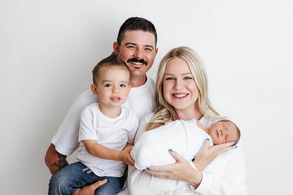 Happy family of four posing together, parents holding toddler son and swaddled newborn baby by Romoland newborn photographer.
