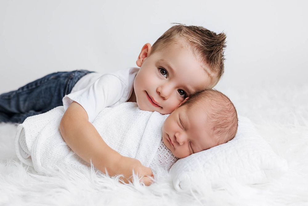 Older toddler boy in white shirt lying on fluffy white blanket cuddling sleeping newborn baby sibling.