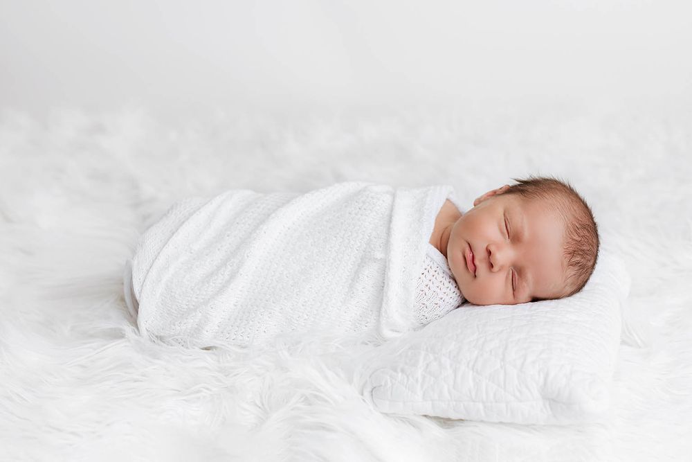 Newborn baby swaddled in white wrap sleeping peacefully on a fluffy white blanket with a knit pillow.