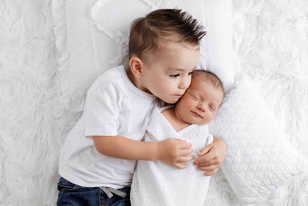 Young boy in white shirt tenderly cuddles sleeping newborn baby sibling by Romoland newborn photographer.