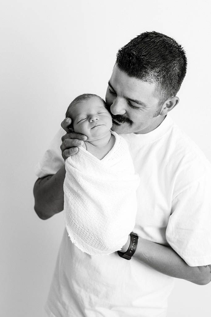 Loving father in white t-shirt tenderly kissing swaddled newborn baby, black and white portrait photo.