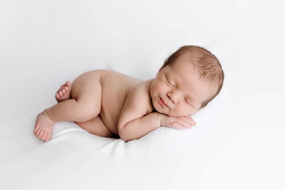 Smiling newborn baby sleeping peacefully on white blanket in classic curled pose for professional newborn photography.