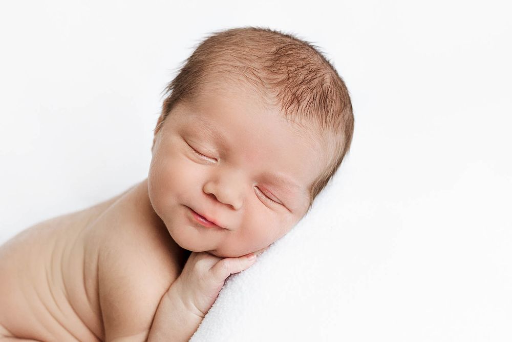 Peaceful newborn baby sleeping and smiling on a soft white blanket in a professional studio photo by Romoland newborn photographer.