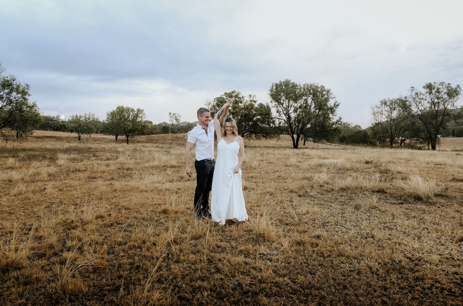 Bride and groom holding hands in golden field with trees under cloudy sky at rustic outdoor wedding.