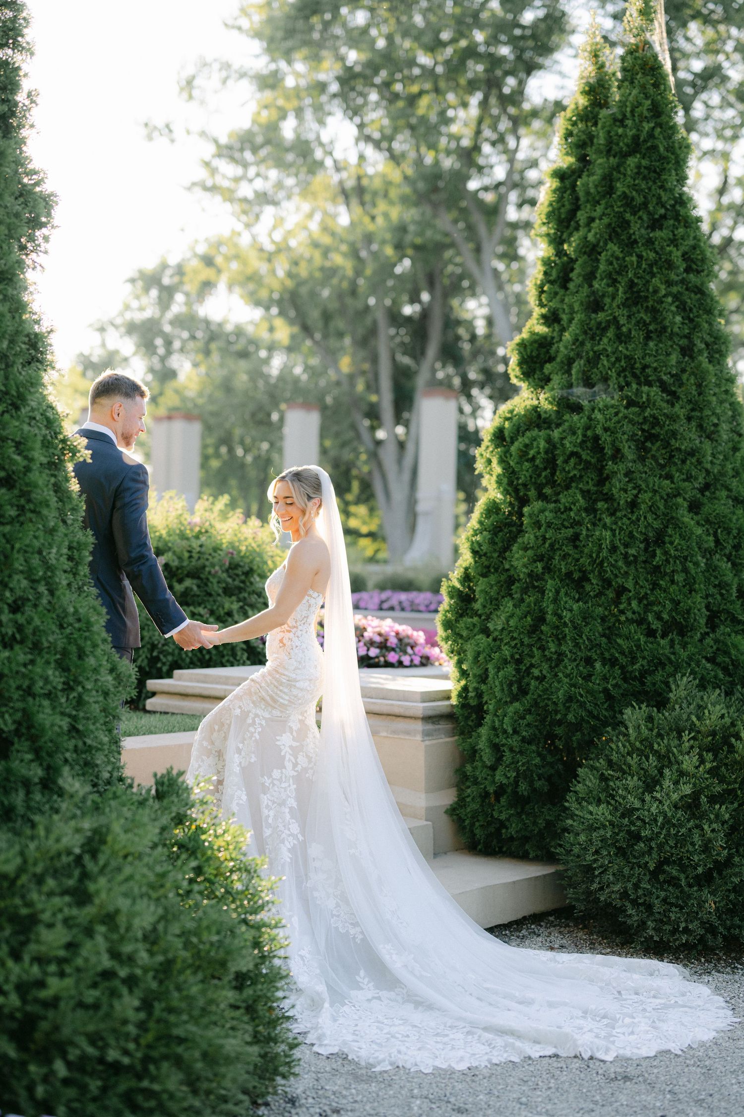 Armour House Wedding - Bride in lace gown with long veil holds groom's hand in elegant garden with tall evergreen topiaries.
