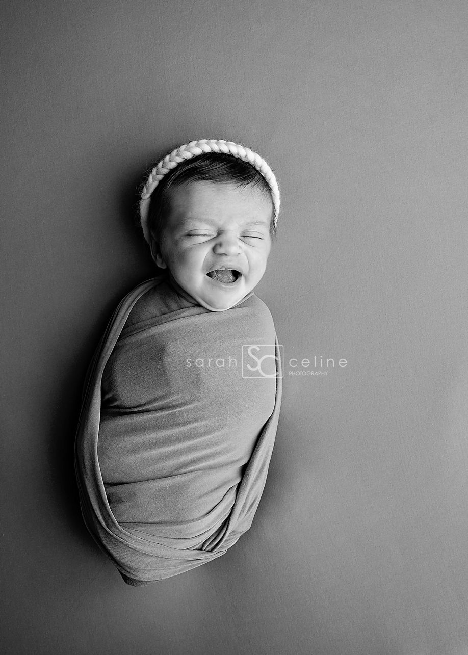 Black and white photo of a swaddled newborn baby smiling, wearing a white knit headband, by Sarah Celine Photography.