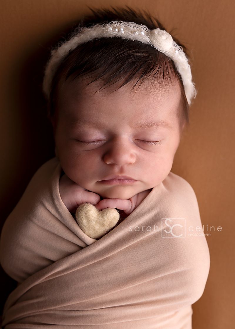 Sleeping newborn baby girl wrapped in beige swaddle holding a heart, wearing a white lace headband on brown backdrop.