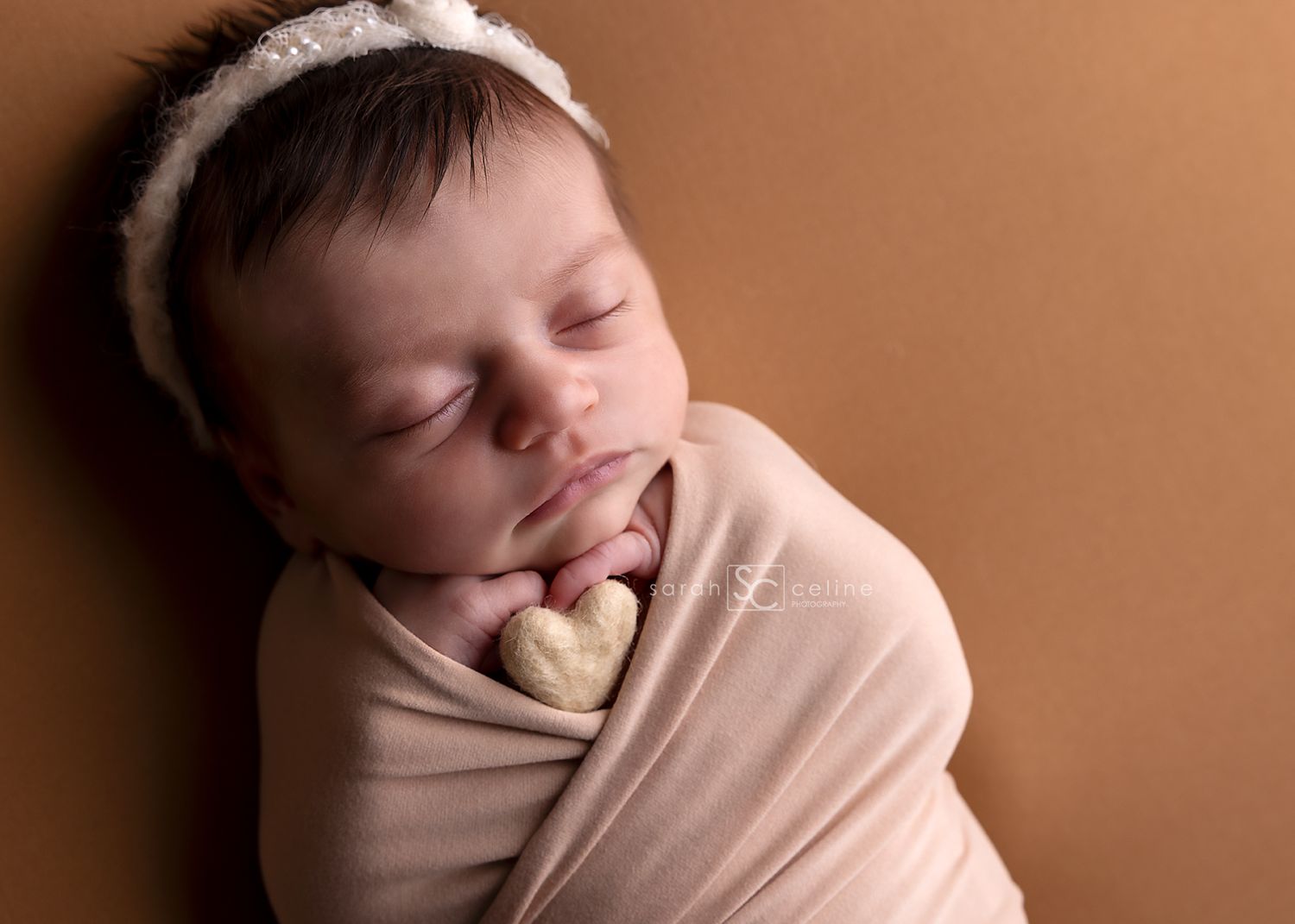 Newborn baby girl swaddled in peach wrap holding a heart prop, wearing a white lace headband on brown backdrop.