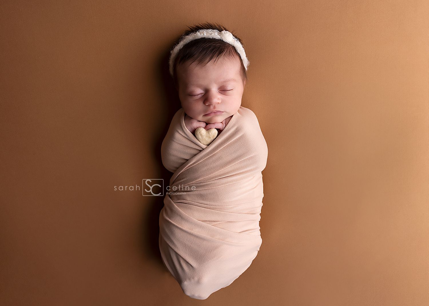 Newborn baby swaddled in a soft pink wrap with a white headband, sleeping peacefully on a warm brown background.