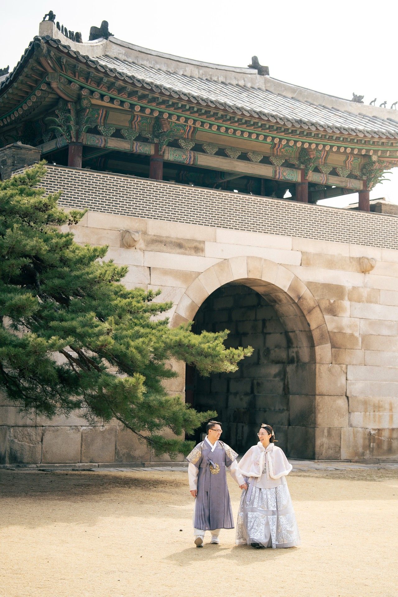 Couple in traditional Korean hanbok attire holding hands before a historic stone gate at a Korean palace.
