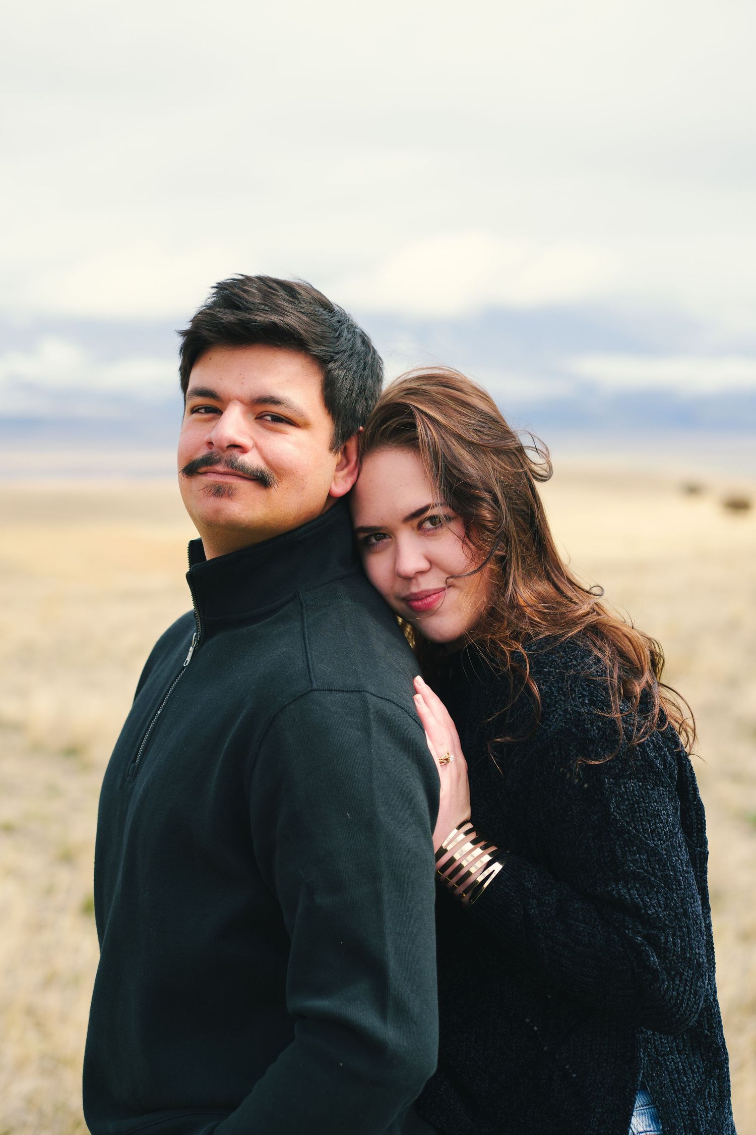 Young couple posing back-to-back in golden prairie field with mountains visible in the distance.