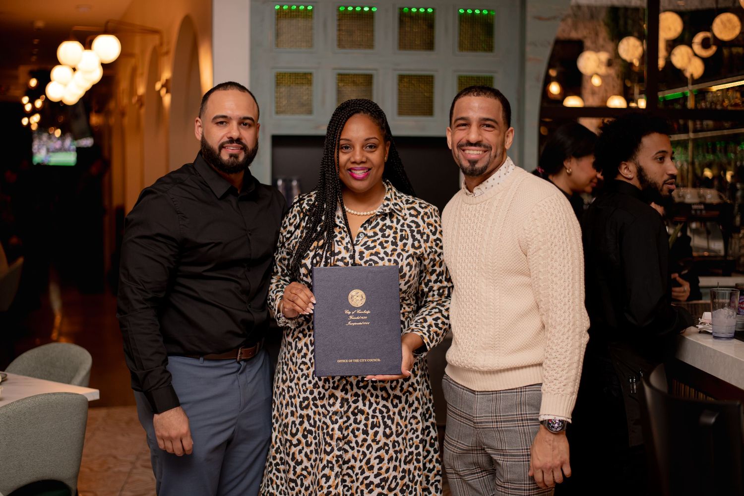 Three people smiling together holding a navy award booklet at an indoor restaurant celebration event.