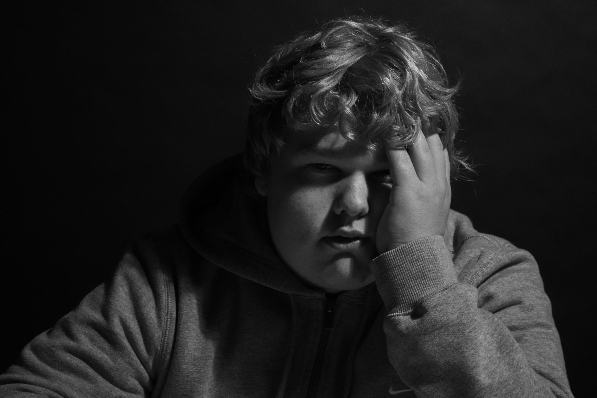 Black and white portrait of a young boy in a hoodie, resting his hand on his face with a pensive expression.