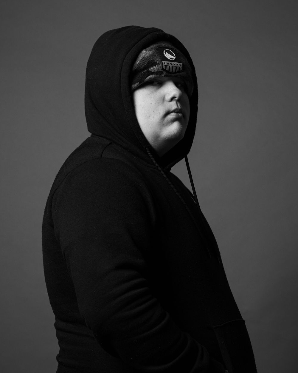Black and white portrait of a person in a black hoodie wearing a sleep mask, posed against a gray background.