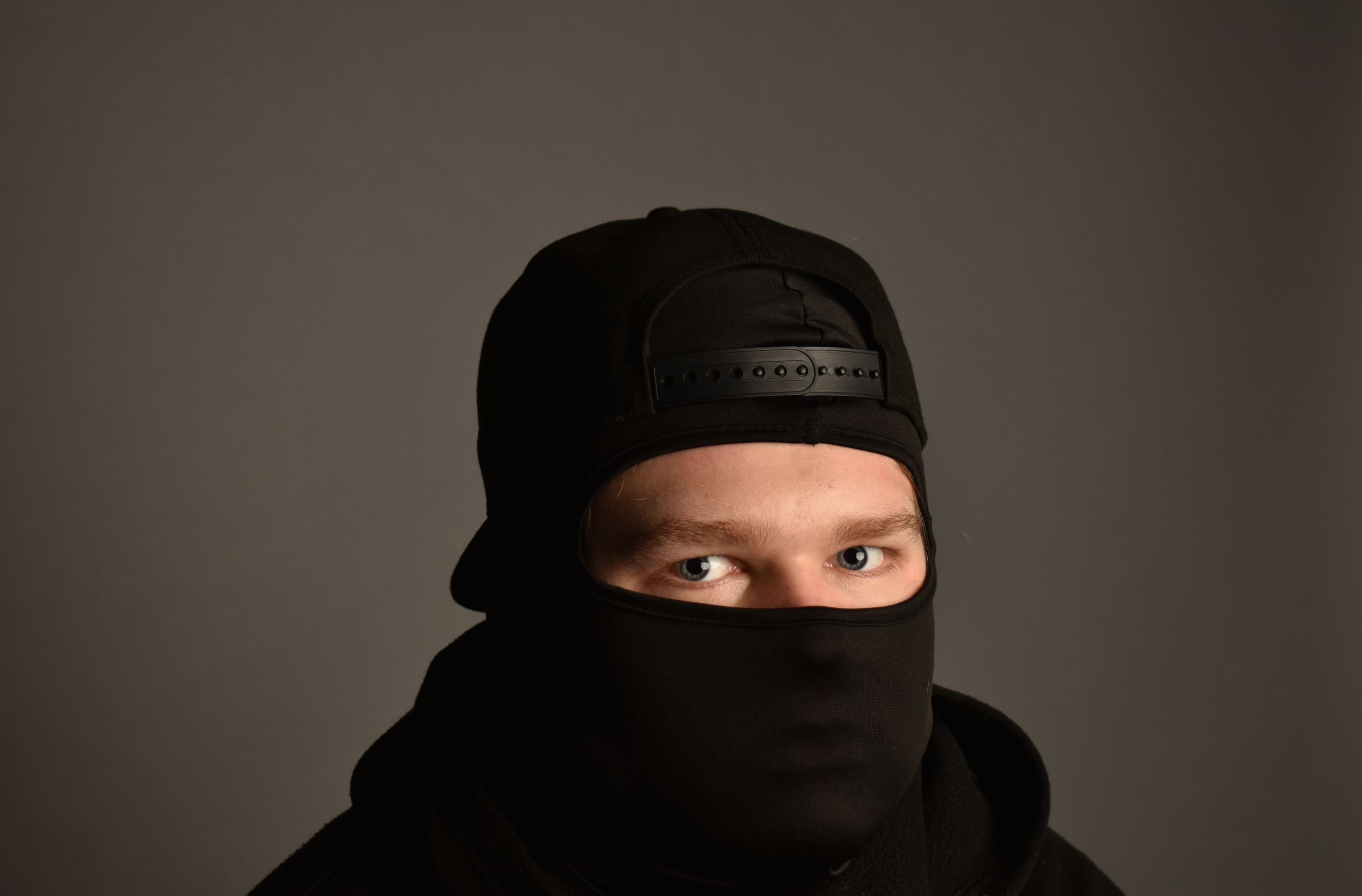 Person wearing black balaclava and snapback cap against gray background, mysterious street style look.