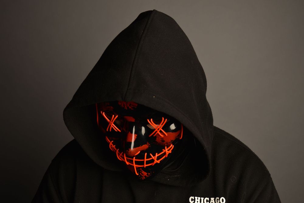Person wearing a black hoodie and glowing red LED purge mask in a dark studio setting.