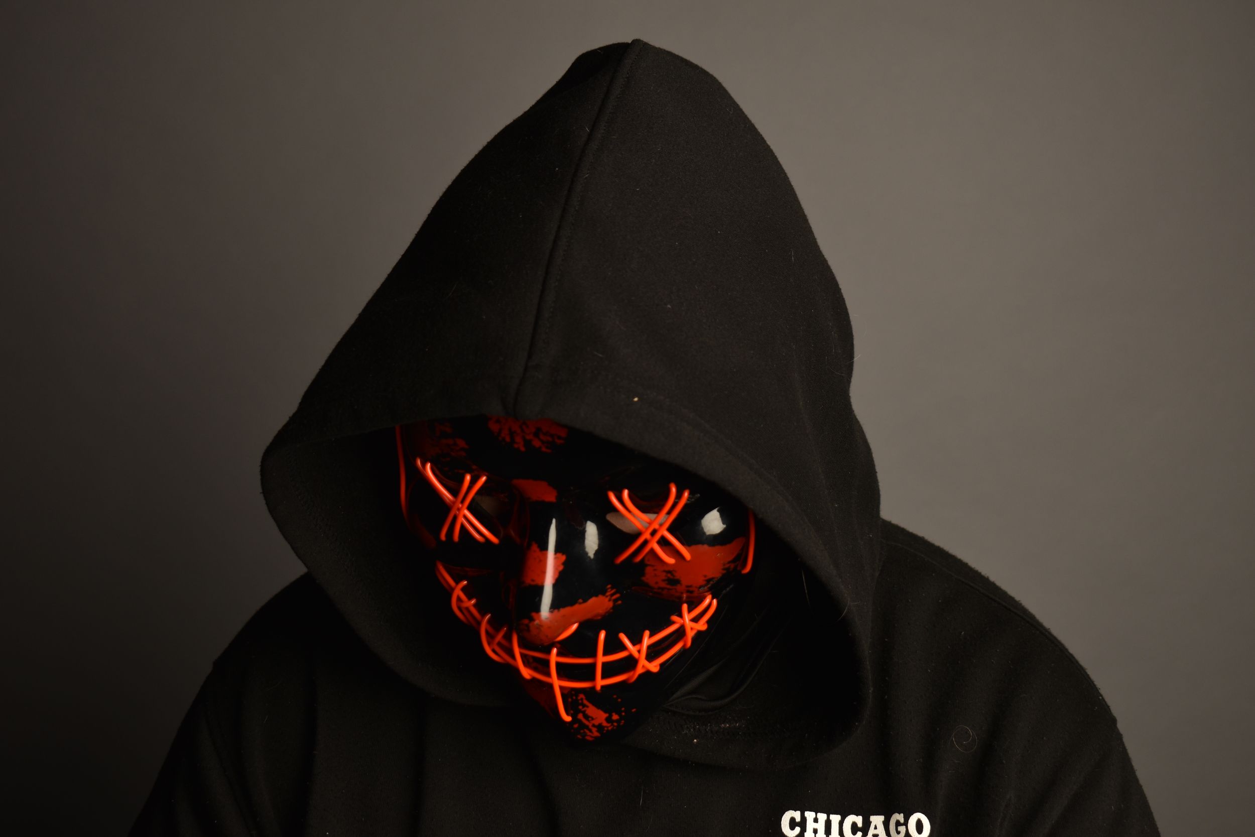 Person wearing a black hoodie and glowing red LED purge mask in a dark studio setting.