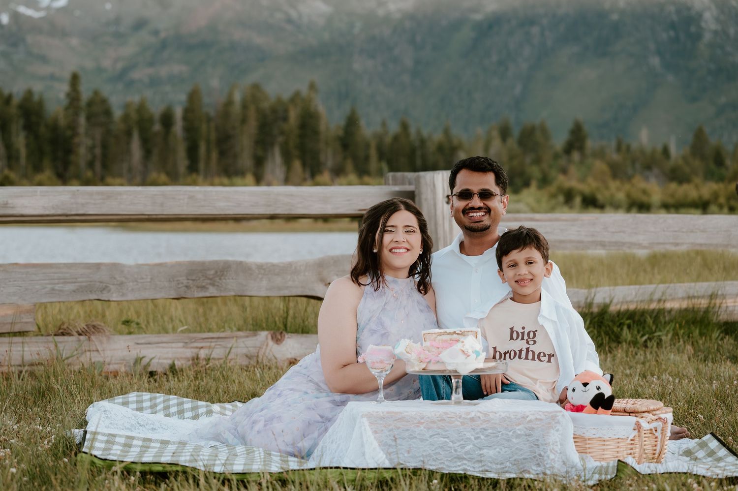 Family of three sitting on a blanket in a meadow, child wearing a big brother shirt, mountains and trees in background.