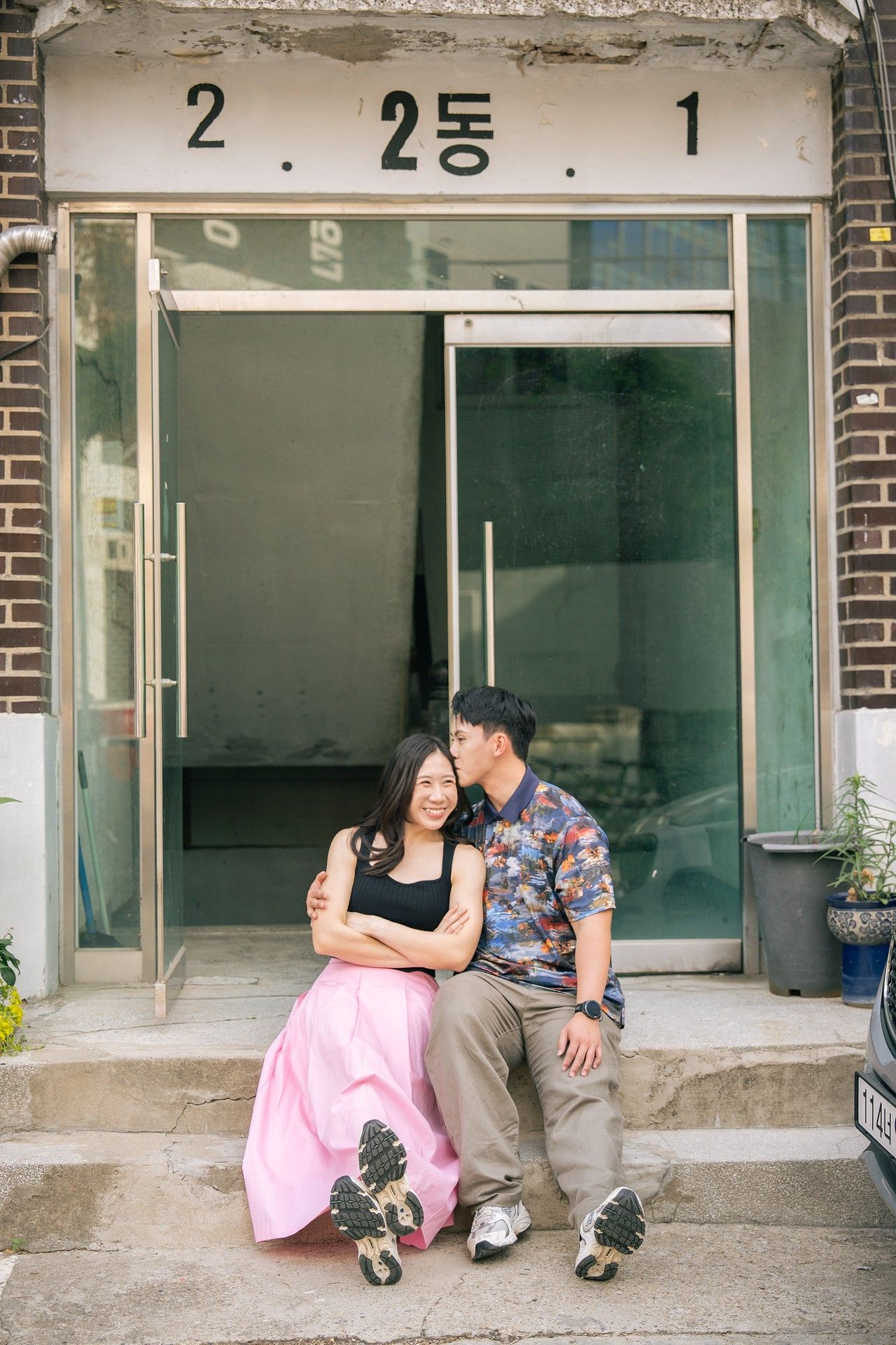 Couple sitting on steps outside a Korean building, man kissing woman's cheek, she wears a pink skirt.