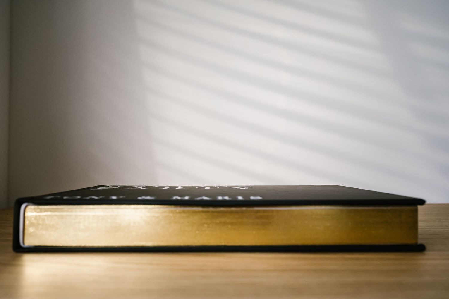A thick gold-edged Bible rests on a wooden table with soft light and shadow patterns on the wall behind it.