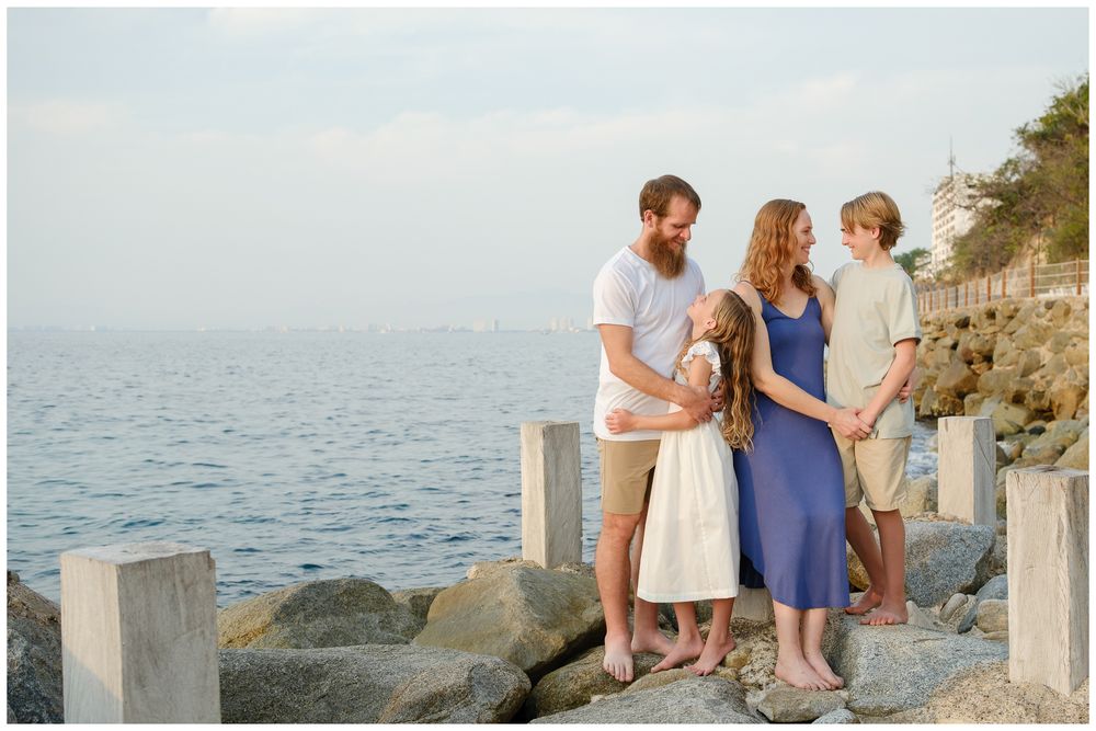 Family of four posing on rocky shoreline at golden hour with calm blue ocean and distant lighthouse in background.