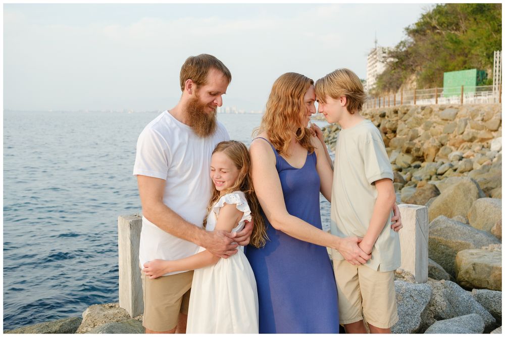Family of four smiling together by rocky seaside, parents embracing children in casual summer attire near ocean.