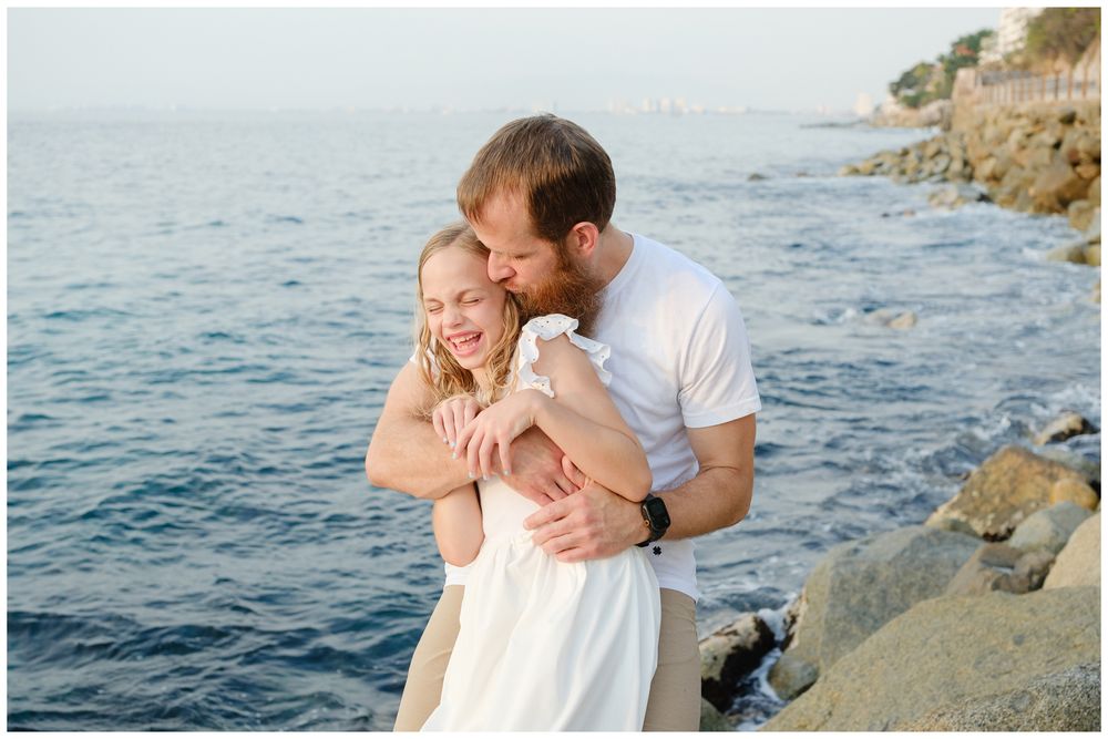 Couple embracing and laughing on rocky seaside shore, man in white shirt hugging woman in white dress at golden hour.