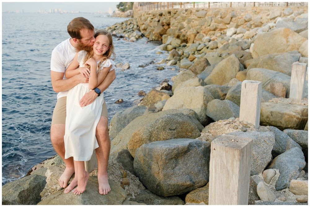 Couple embracing on rocky shoreline at sunset, man kissing woman in white dress near calm blue ocean.