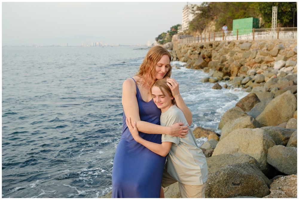 Two women embracing warmly on a rocky seaside shore, wearing casual summer outfits with ocean waves in the background.