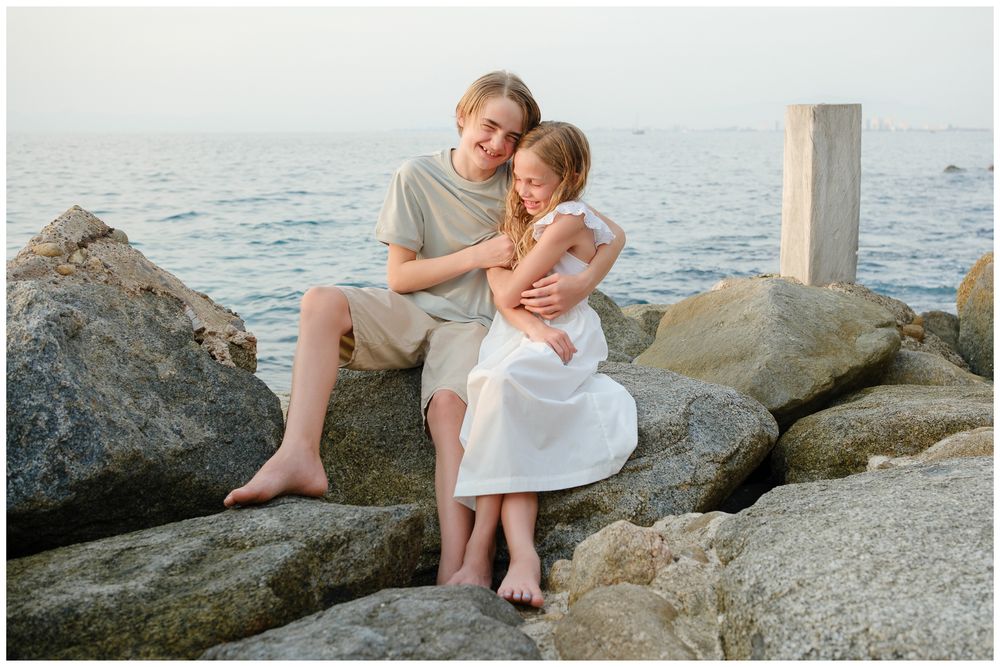 Boy and girl sitting on rocks by the sea, sharing a sweet embrace during golden hour photography.