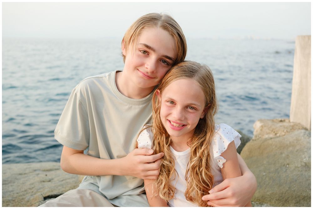 Two siblings smile together on rocky seaside, boy hugging younger girl holding ice cream cone.