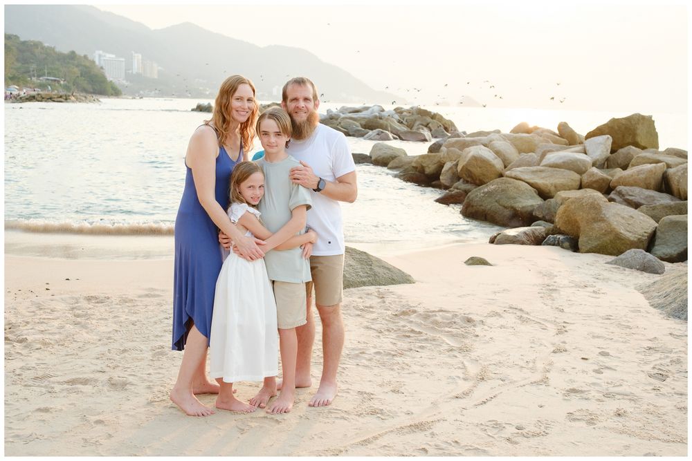 Happy family of four poses on sandy beach at golden hour with rocky shoreline backdrop.