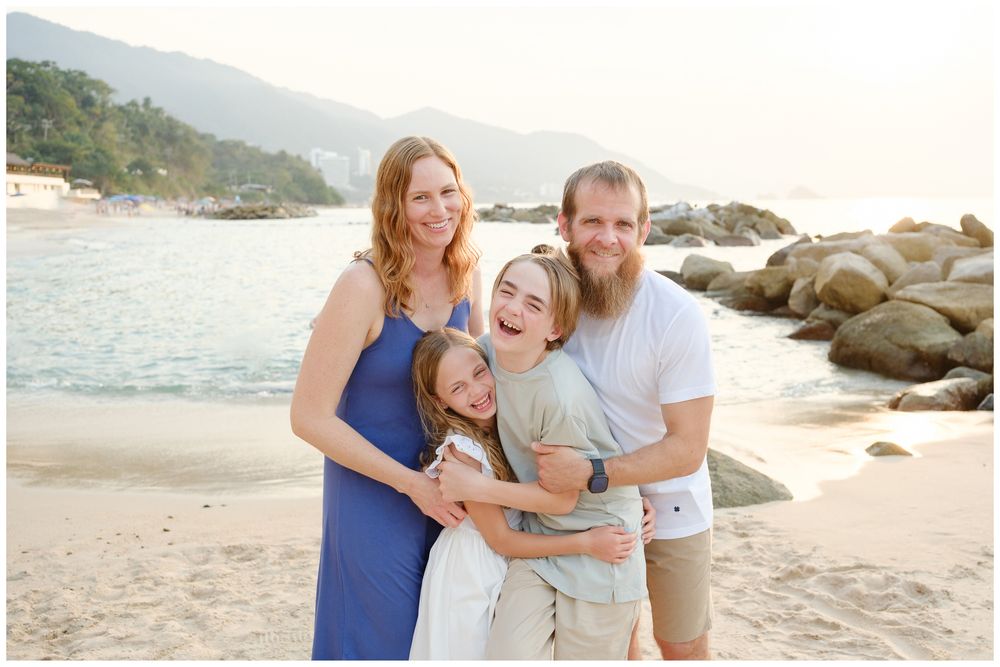 Joyful family laughing together on tropical beach, parents embracing two children near rocky coast.