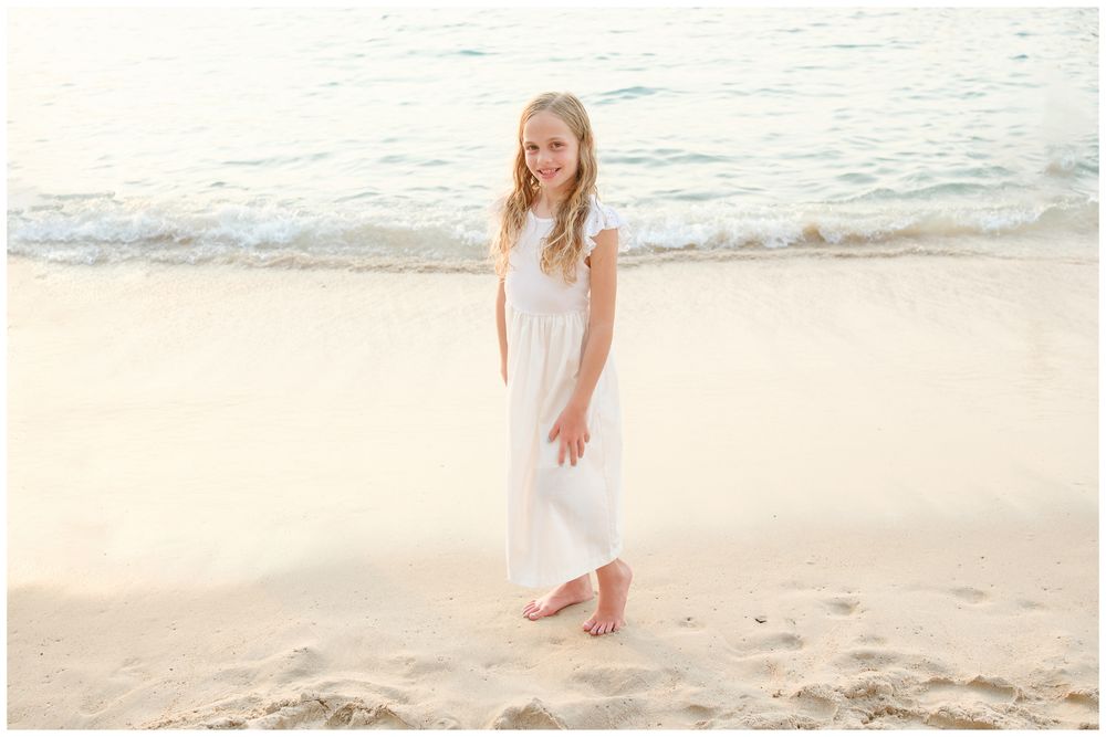 A smiling young girl in a white maxi dress stands barefoot on a sandy beach with gentle ocean waves behind her.
