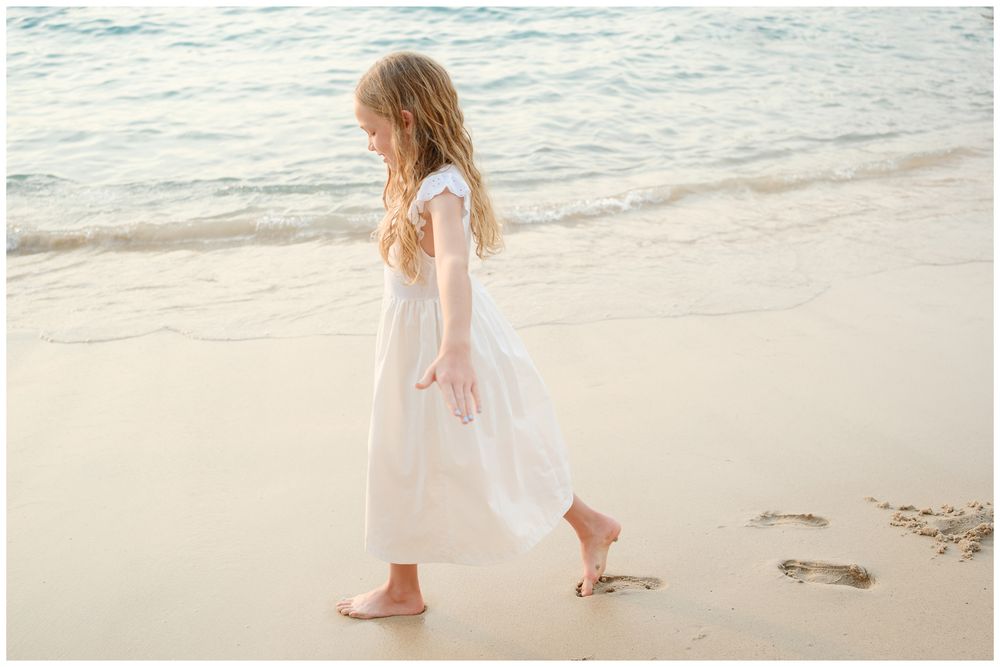 A young girl in a white dress walks barefoot along a sandy beach with gentle ocean waves in the background.